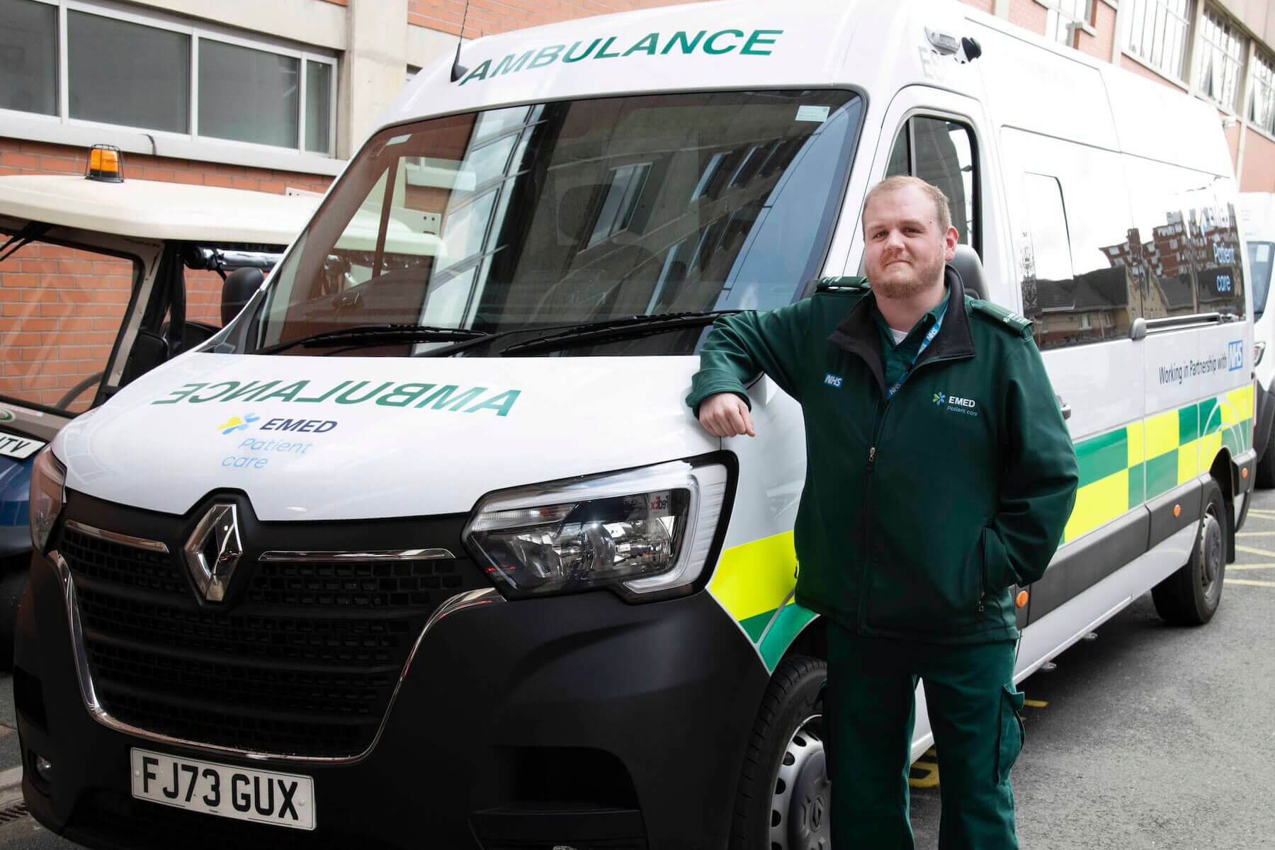 EMED Patient Care Renal Manager Steve Lovell standing in front of a branded Patient Care ambulance, smiling, wearing a green uniform with the EMED Patient Care and NHS logos visible on the jacket and van.