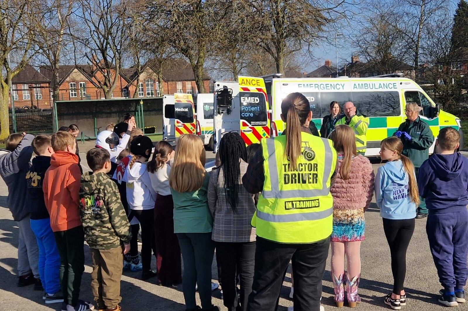 A group of school children gathers outdoors around EMED Patient Care staff and ambulance during a community education event. The children, dressed in casual clothing, are attentively listening. The EMED team members are demonstrating equipment, engaging with the students, and showcasing the ambulances. The background features trees, a school building, and clear skies.